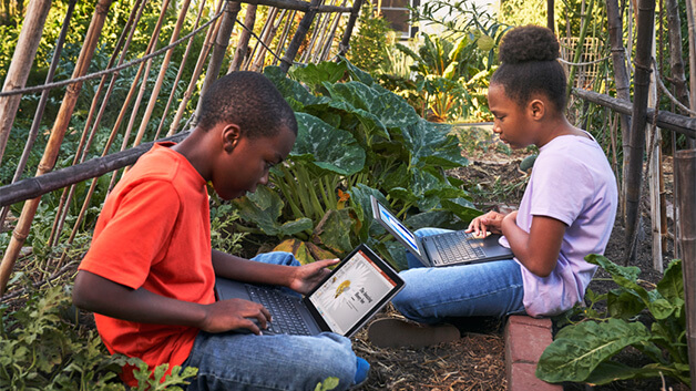 Two K-12 students, male and female, sitting in community garden. Both students are using laptops.